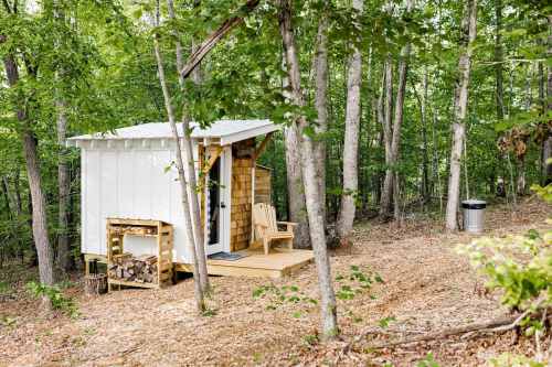 A small cabin in a wooded area, featuring a wooden deck, firewood stack, and a chair outside.