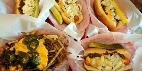 A variety of hot dogs and loaded fries topped with cheese, jalapeños, and pickles, served in paper wrappers.