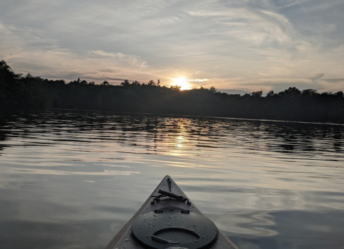 A kayak on calm water at sunset, with trees silhouetted against a colorful sky.