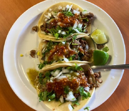 Three tacos filled with meat, topped with onions, cilantro, and sauces, served with lime wedges on a white plate.