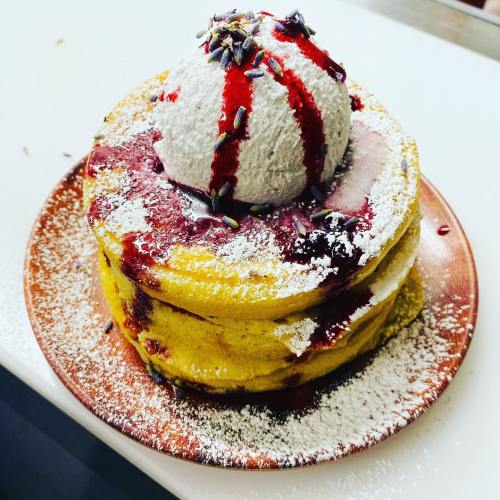 Stack of fluffy pancakes topped with a scoop of ice cream, berry sauce, and a sprinkle of lavender, dusted with powdered sugar.