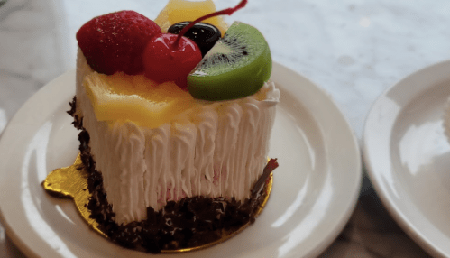 A slice of cake topped with strawberries, cherries, kiwi, and pineapple, resting on a white plate.
