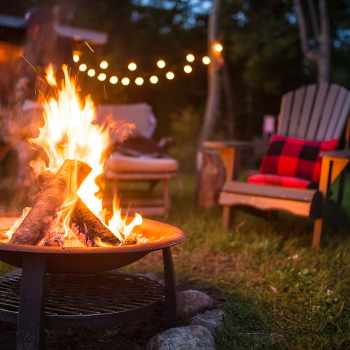A cozy campfire burns brightly in a backyard, surrounded by chairs and string lights in the evening.