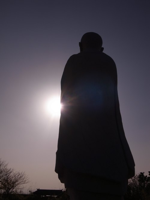 Silhouette of a statue against a bright sun, with a clear sky and trees in the background.
