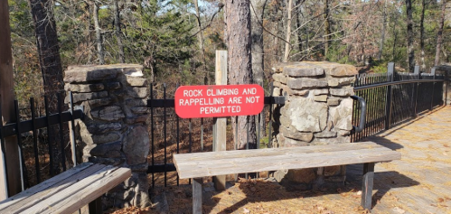 A wooden bench near a sign stating "Rock climbing and rappelling are not permitted" in a wooded area.