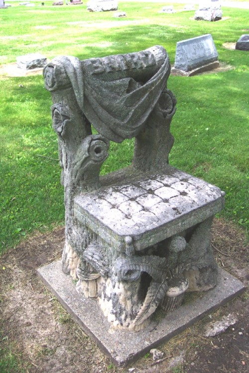 A weathered stone chair with intricate carvings, set on grass in a cemetery.