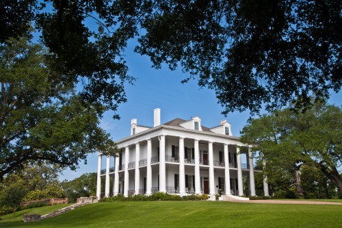 A grand white mansion with columns, surrounded by lush greenery and a clear blue sky.