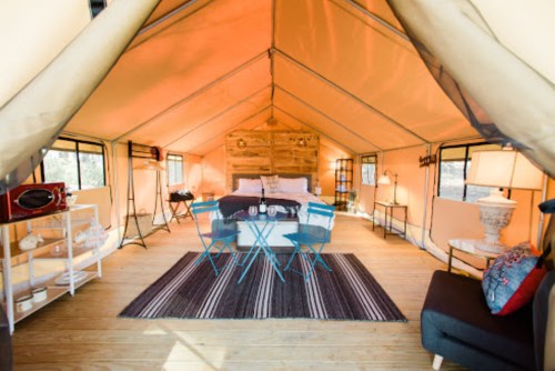 Cozy glamping tent interior with a bed, dining area, and stylish decor, featuring warm lighting and wooden accents.