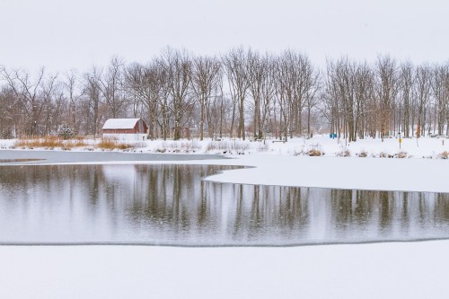 A snowy landscape featuring a calm pond, bare trees, and a red barn in the background.