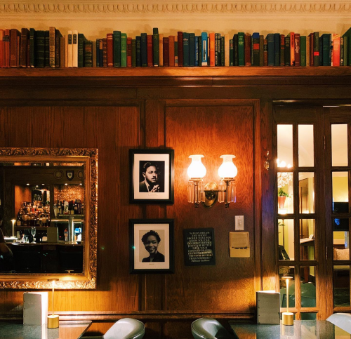A cozy bar interior featuring a wooden wall, books on a shelf, framed portraits, and warm lighting.