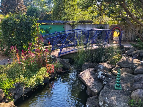 A blue bridge over a pond surrounded by lush greenery and colorful flowers in a serene garden setting.