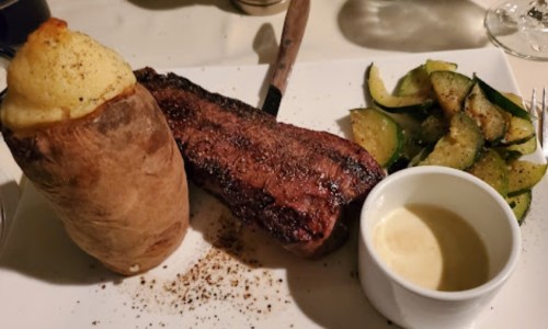 A plate featuring a baked potato, grilled steak, sautéed zucchini, and a small dish of sauce.