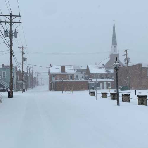 A snowy street scene with a church steeple in the background, power lines, and buildings partially obscured by falling snow.