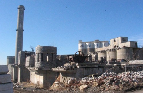 Abandoned industrial site with a tall smokestack, crumbling structures, and scattered debris under a clear blue sky.