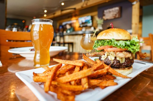 A juicy burger with lettuce, tomato, and bacon, served with sweet potato fries and a glass of beer in a cozy restaurant.