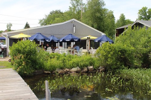 A cozy restaurant by a pond, featuring outdoor seating under blue umbrellas and surrounded by greenery.