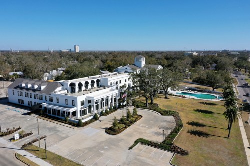 Aerial view of a large white building with palm trees, a pool, and a spacious parking area on a clear day.