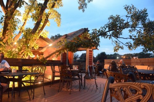 Outdoor dining area with wooden tables and chairs, surrounded by trees and soft evening lighting.