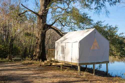 A white canvas tent cabin on stilts by a calm river, surrounded by trees and a picnic table nearby.