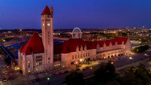 Aerial view of a historic train station at dusk, featuring a tall clock tower and illuminated buildings against a city skyline.