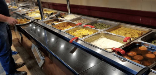 A buffet with various dishes in metal trays, including rice, vegetables, and sauces, with a person serving food.