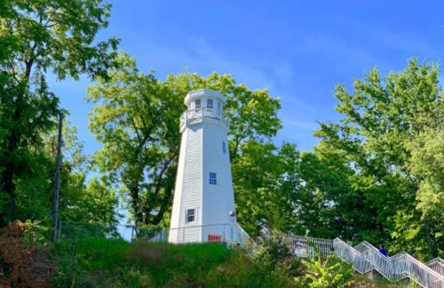 A white lighthouse surrounded by lush green trees under a clear blue sky.