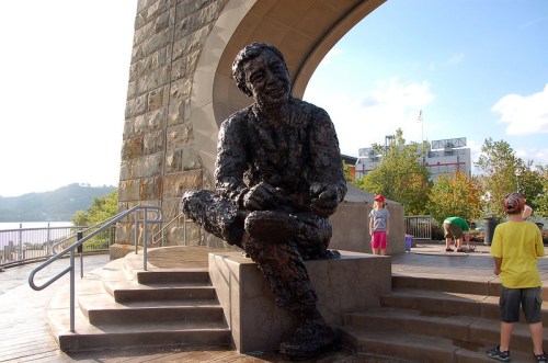 A bronze statue of a seated man, surrounded by trees and children playing, with a river and buildings in the background.