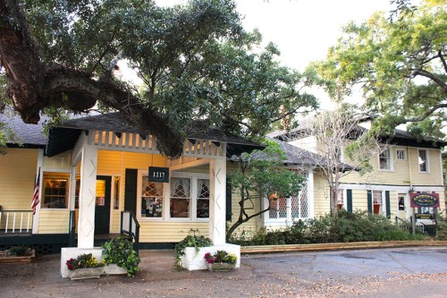 A charming yellow building with a porch, surrounded by trees and flowers, featuring a welcoming entrance.