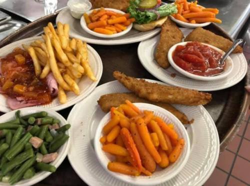 A tray with various dishes: fried fish, ham with sauce, fries, green beans, and carrots, served with salad and dipping sauce.