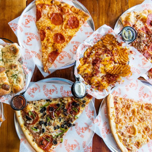 A variety of pizzas, including pepperoni and veggie, alongside loaded fries and garlic knots on a wooden table.