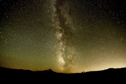 A stunning night sky filled with stars and the Milky Way, silhouetted by distant mountains and a soft glow on the horizon.