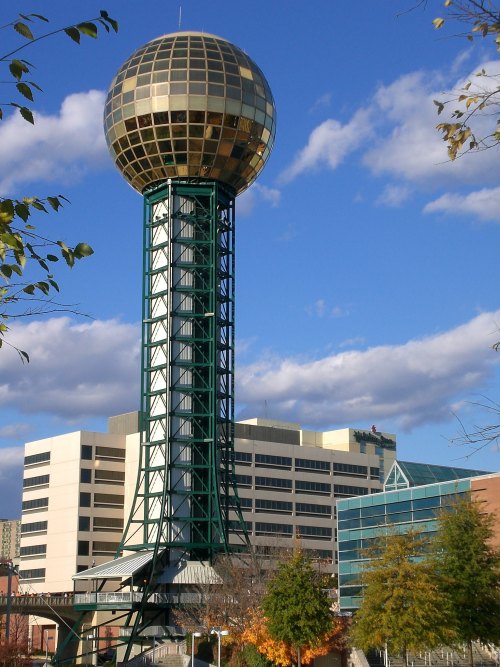 A tall observation tower with a golden dome, surrounded by buildings and trees under a blue sky with clouds.
