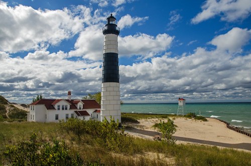 A tall lighthouse beside a sandy beach, with a white and black striped tower under a cloudy sky and blue water.
