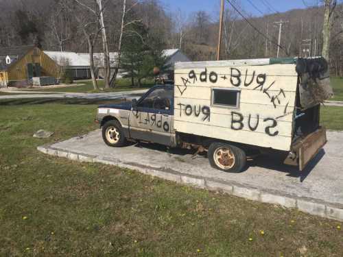 A weathered truck with a wooden structure, painted with "Bugle Tour Bus," parked on a concrete slab in a rural area.