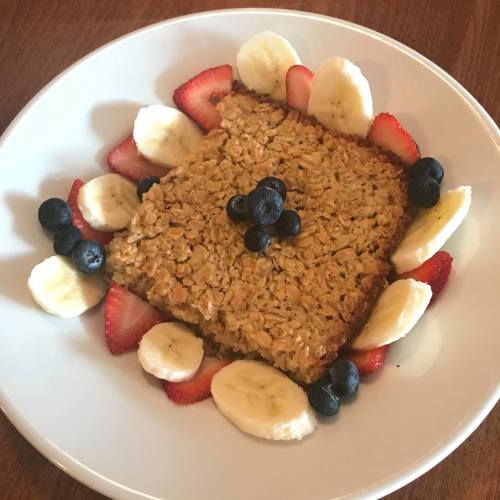 A square baked oatmeal topped with blueberries, surrounded by slices of banana and strawberries on a white plate.