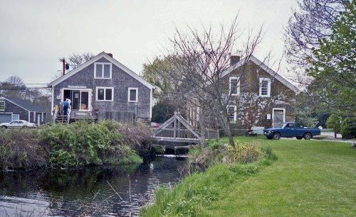 A serene scene of two houses by a pond, with a bridge and a truck parked on the grassy bank.