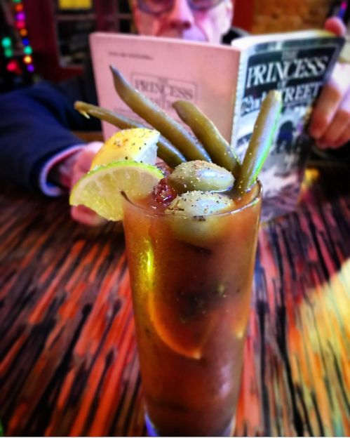A close-up of a Bloody Mary garnished with green beans and a lime slice, with a person reading in the background.