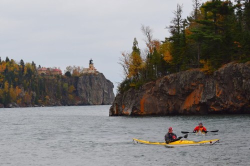 Two kayakers paddle in a calm lake, with a lighthouse and autumn trees in the background.