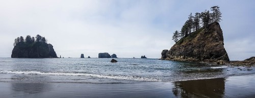 A panoramic view of a rocky beach with islands and trees under a cloudy sky. Waves gently lap at the shore.