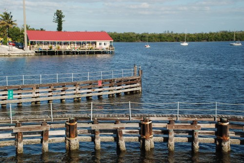 A wooden pier extends into a calm body of water, with a red-roofed building and boats in the background.