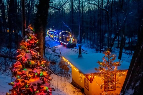 A snowy scene at dusk featuring a brightly lit train and decorated trees near a cozy cabin.
