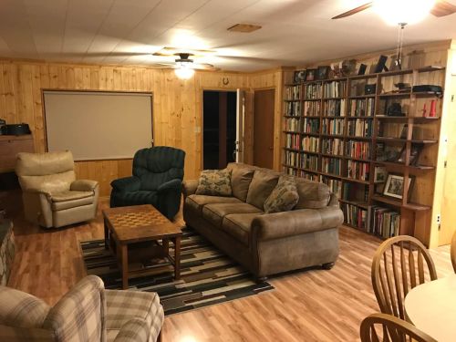 Cozy living room with wooden walls, a sofa, armchairs, a coffee table, and a bookshelf filled with books.