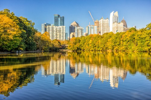 City skyline reflecting in a calm lake, surrounded by vibrant autumn foliage and clear blue skies.
