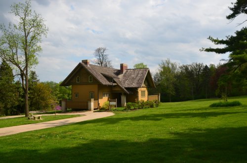 A yellow house with a sloped roof surrounded by green grass and trees under a partly cloudy sky.
