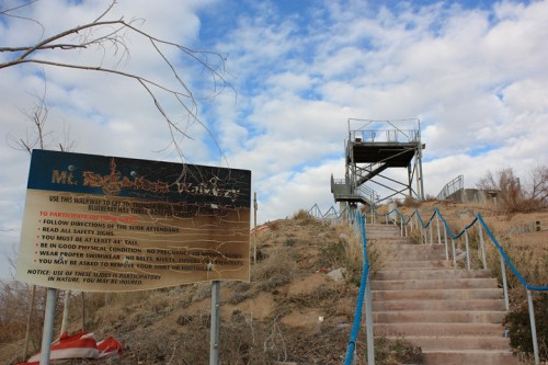 A sign at the base of stairs leading to an observation platform on a hill, with cloudy skies above.