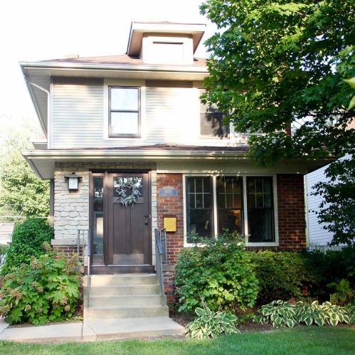 A two-story house with a brick and stone facade, surrounded by greenery and featuring a front porch and steps.