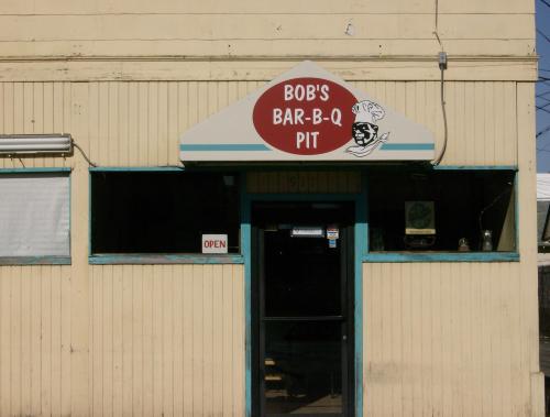 Sign for "Bob's Bar-B-Q Pit" above the entrance of a rustic building, with an open sign visible.