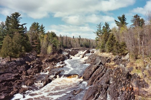 A scenic river flows through rocky terrain, surrounded by lush trees and a blue sky with scattered clouds.