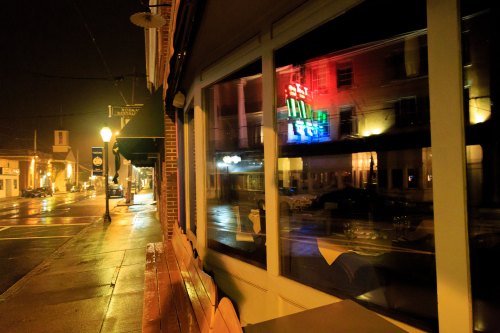 A nighttime street scene with a restaurant window reflecting colorful neon lights and wet pavement.