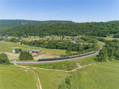 A train curves through a lush green landscape, with hills and a few houses in the background under a clear blue sky.